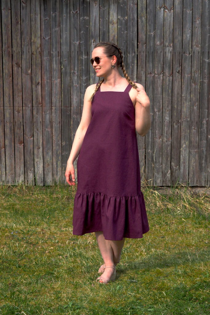 Kuvassa hymyilevä nainen aurinkolasit päässä seisoo ruohikolla, päällään hihaton frillahelmainen pellavamekko.