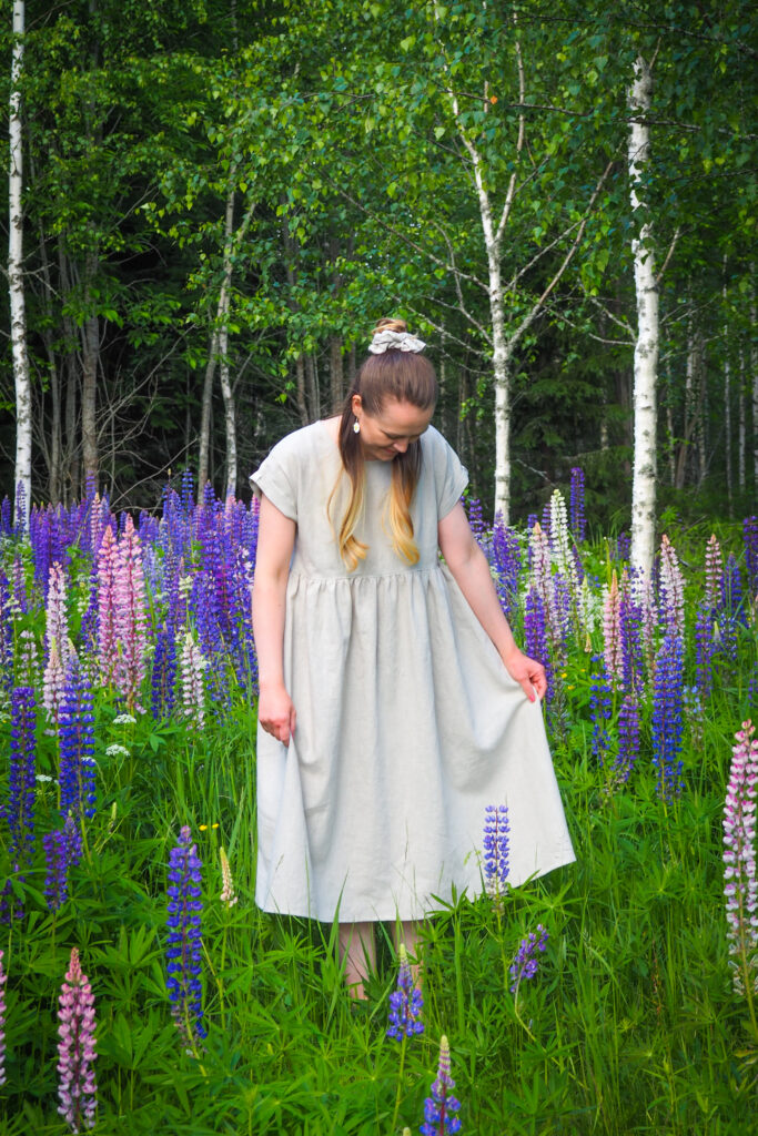 Kuvassa naishenkilö seisoo lupiinien keskellä ja katsoo alaspäin vaalean sävyistä pellavamekkoaan. Hiukset hänellä on osaksi solmittu nutturalle hiusdonitsilla.