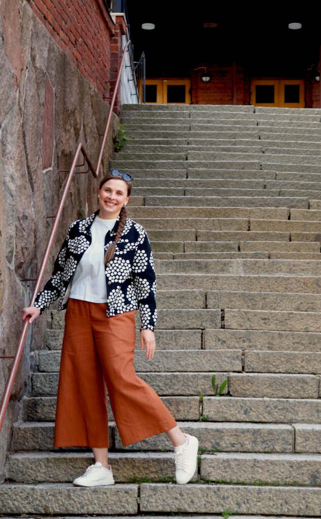 Kuvassa naishenkilöllä päällä kuviollinen takki ja kanelin sävyiset culottes-pellavahousut, hän poseeraa kiviportailla ja pitää kaiteesta kiinni, katse kameraan ja hymyilee.