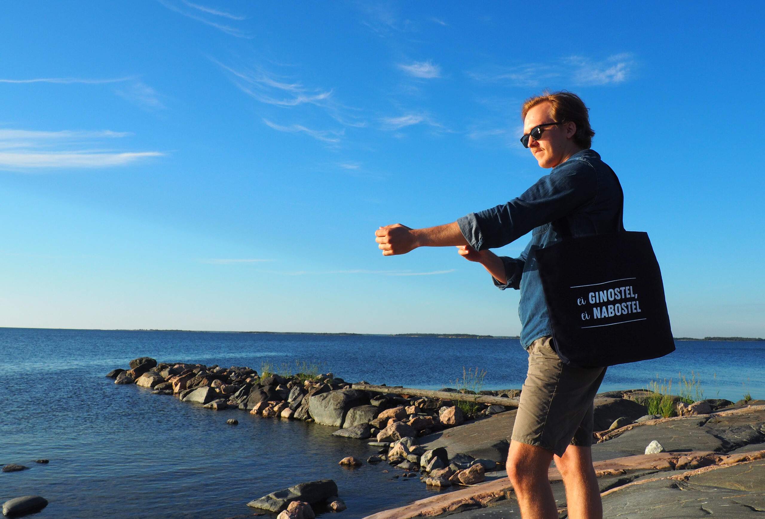Kuvassa farkkupaitaan ja shortseihin pukeutunut mies käärii hihaansa merenrannalla. Hän seisoo kalliolla ja olallaan hänellä on kangaskassi jossa teksti ei ginostel, ei nabostel.