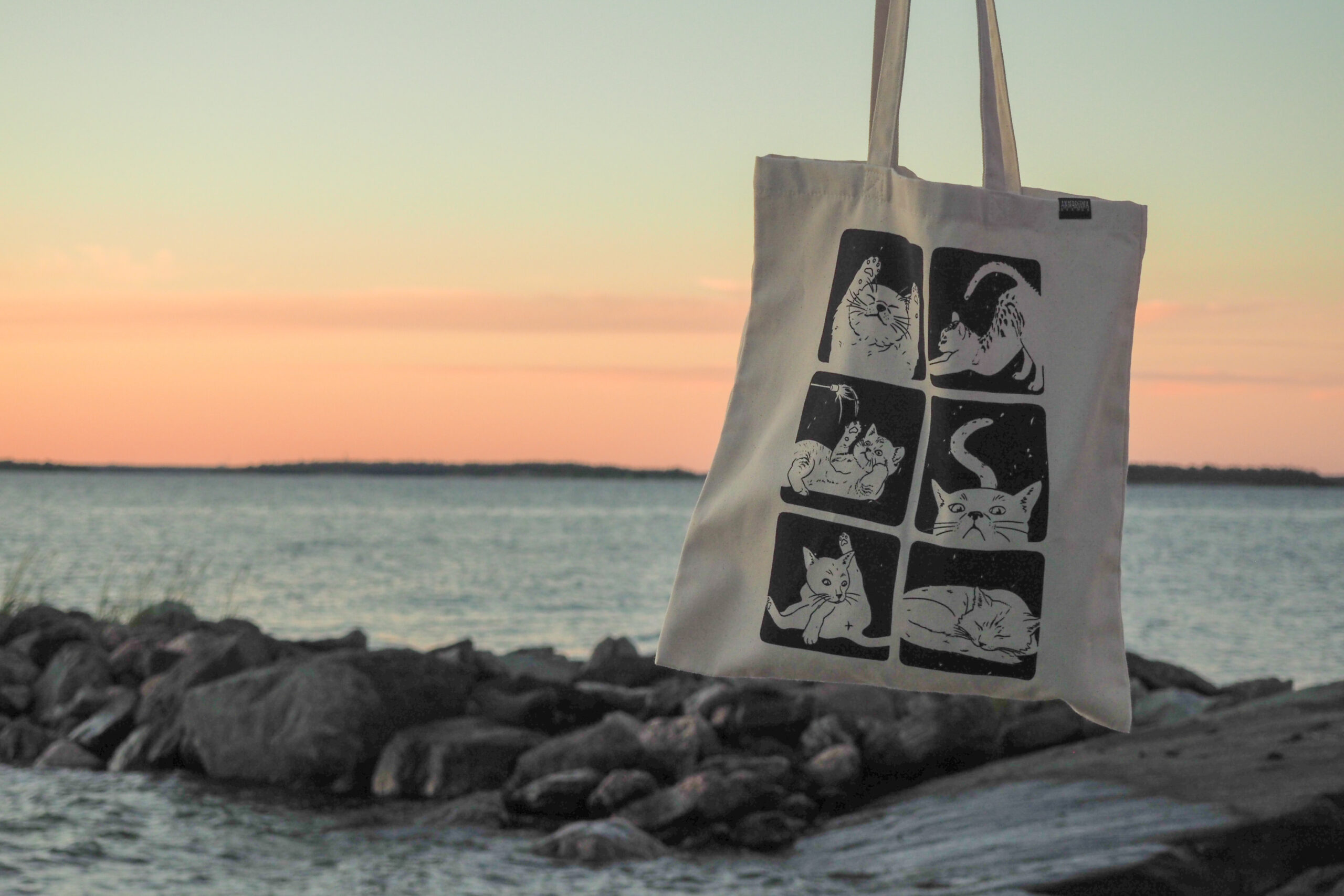 Kuvassa roikkuu merellistä auringonlaskua vasten vaalea kangaskassi jossa on kissa-aiheinen kuvitus.
