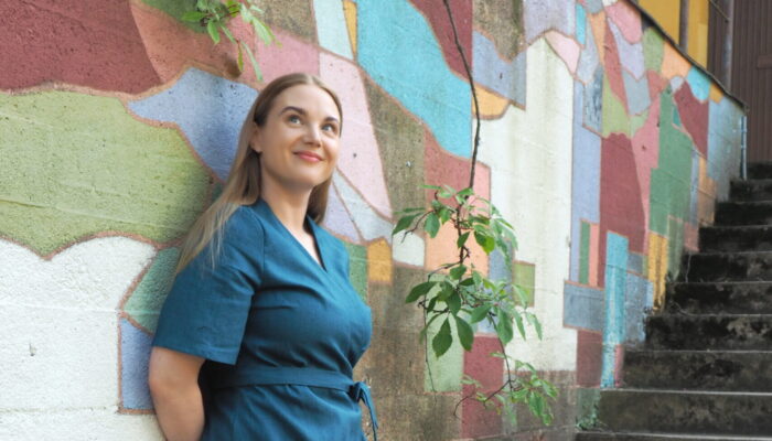 Lilja-mekko Kuvassa hymyilevä nainen nojaa värikästä seinää vasten ja katsoo hiukan yläviistoon. Päällä naisella petroolin värinen hihallinen kietaisumekko. Mekon materiaali pellava.