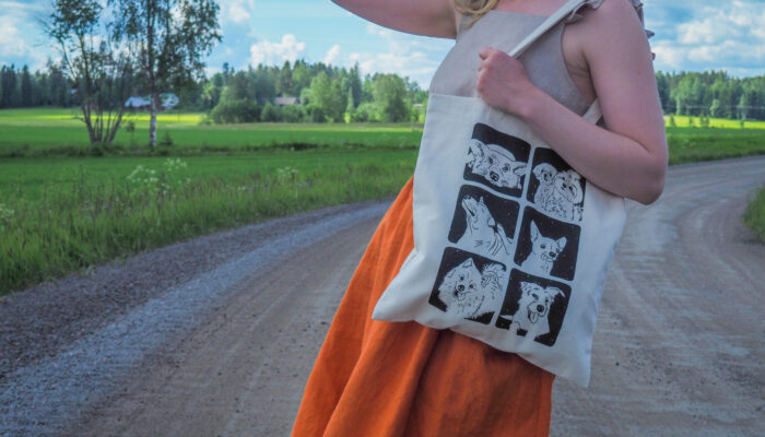 Koira-kangaskassi Lähikuvassa naishenkilö joka seisoo maantiellä päällään oranssi pellavahame ja olalla vaalea koira-aiheinen kangaskassi.