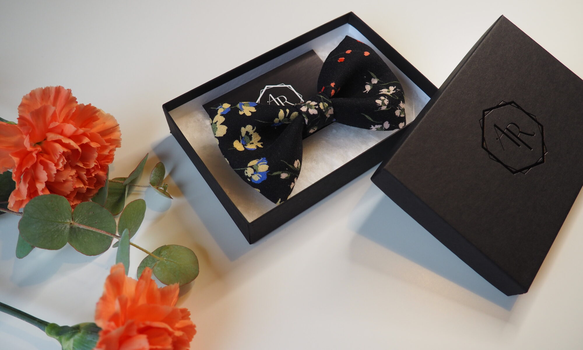 Käsintehty kukkakuvioinen musta rusetti lahjarasiassa oranssien neilikoiden vieressä.