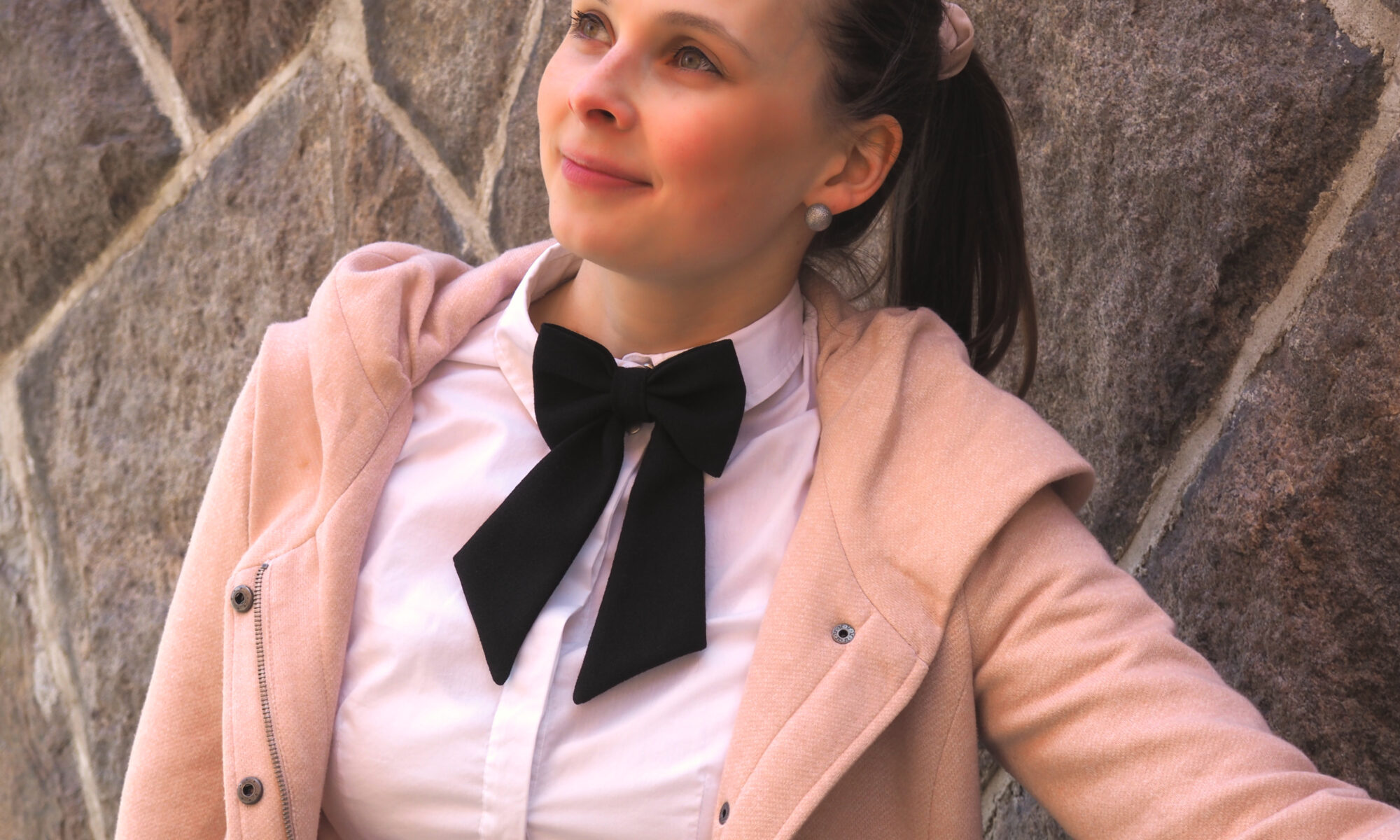 Kuvassa nainen päällään roosan värinen villakangastakki ja valkoinen kauluspaita, kaulassa musta rusetti.