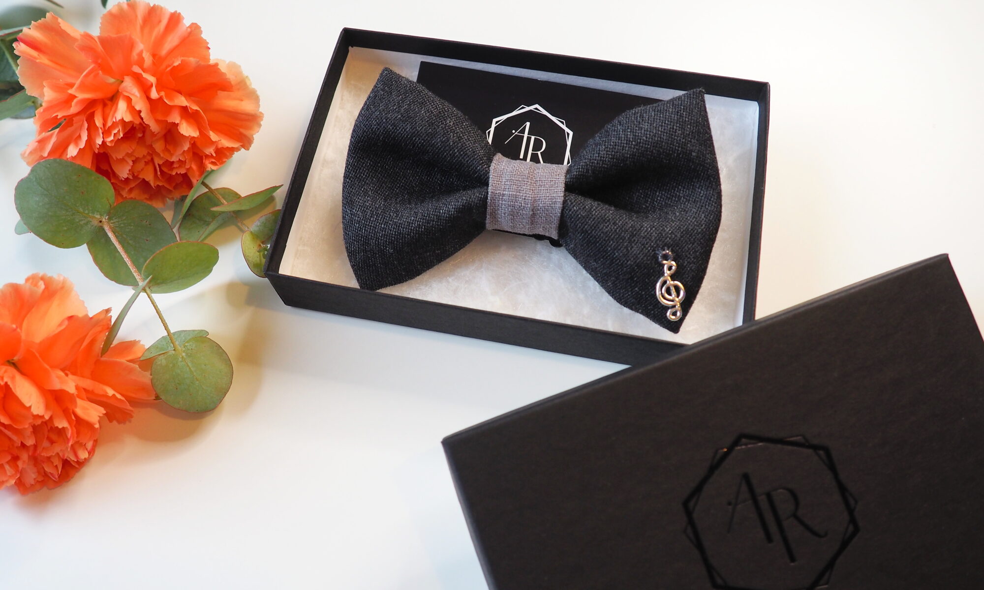 Käsintehty harmaa villarusetti, jossa metallinen nuottiavain ommeltuna rusettiin. Rusetti on lahjarasiassa oranssien neilikoiden vieressä.