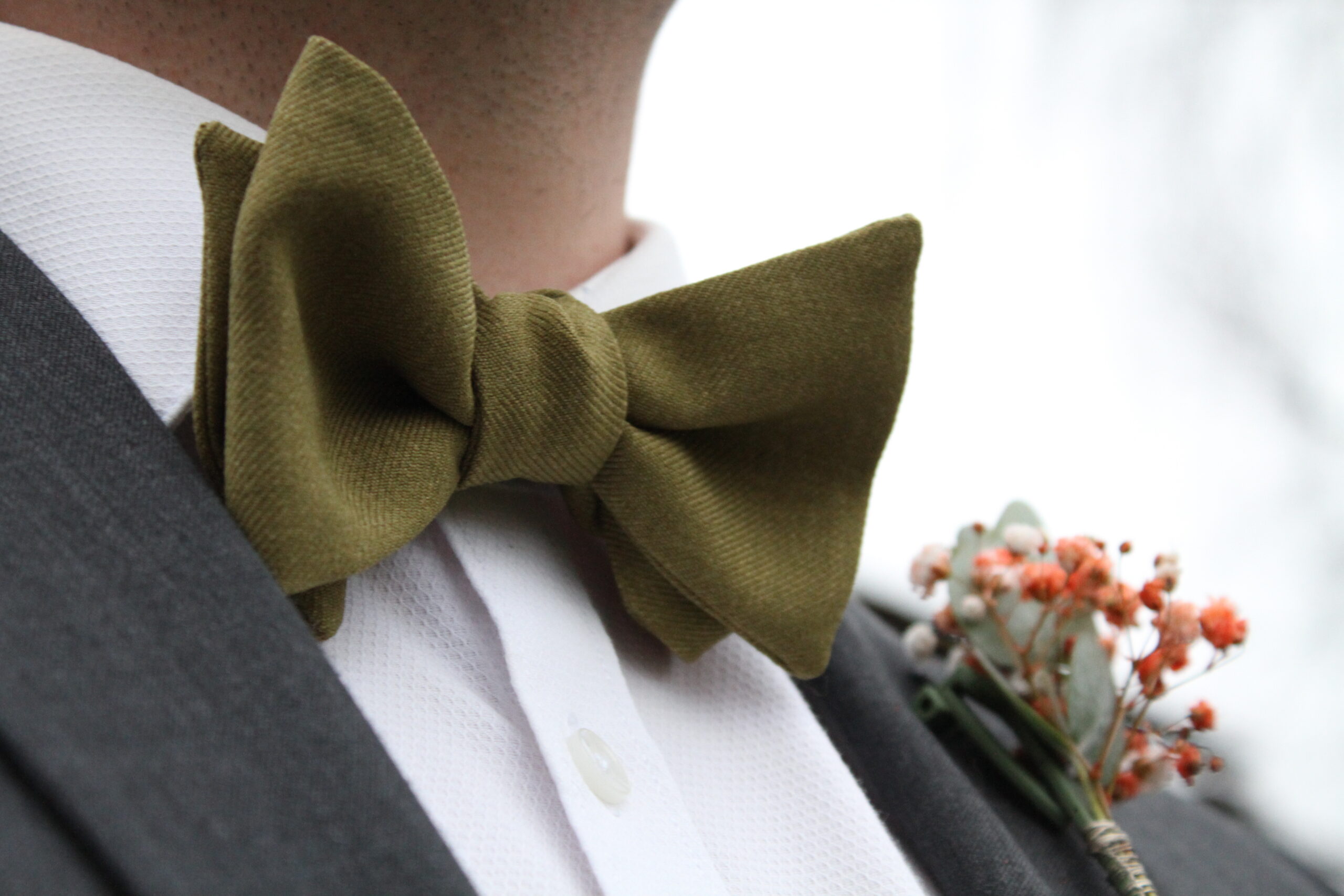 Lähikuva vihreästä rusetista miehen kaulassa. Kuvassa näkyy harmaa puvun takki ja rintapieleen kiinnitetty kukkaviehe.