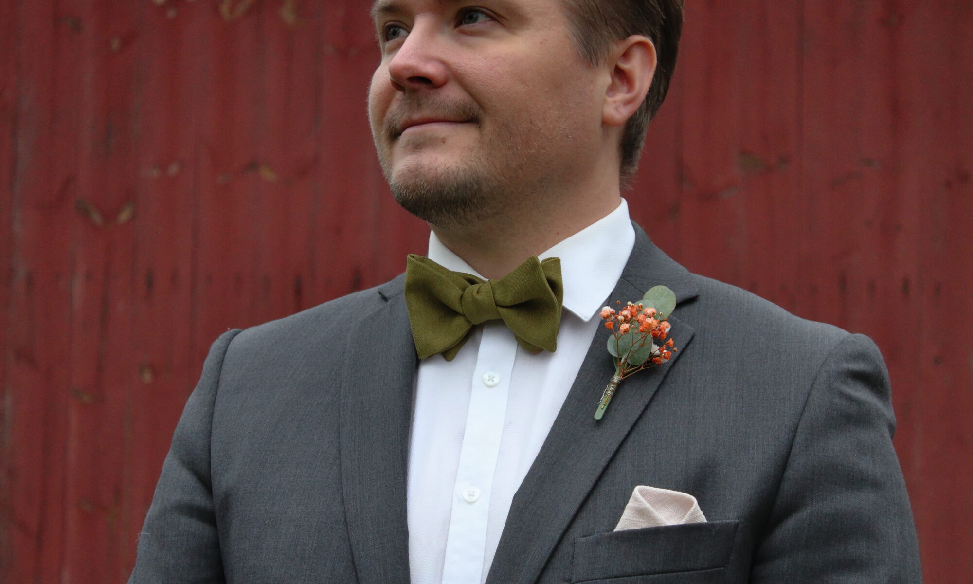 Mies harmaassa puvussa ja valkoisessa kauluspaidassa seisoo punaisen taustan edessä. Hänellä on kaulassa vihreä rusetti ja rintapielessä pieni kukkaviehe.