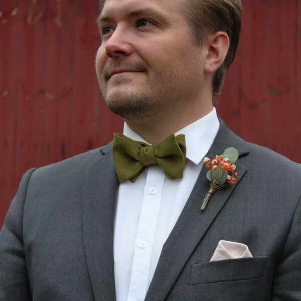 Mies harmaassa puvussa ja valkoisessa kauluspaidassa seisoo punaisen taustan edessä. Hänellä on kaulassa vihreä rusetti ja rintapielessä pieni kukkaviehe.