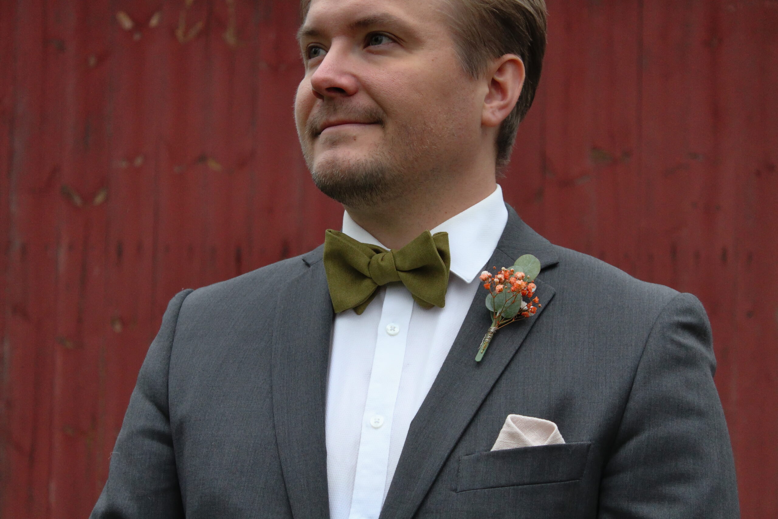 Mies harmaassa puvussa ja valkoisessa kauluspaidassa seisoo punaisen taustan edessä. Hänellä on kaulassa vihreä rusetti ja rintapielessä pieni kukkaviehe.