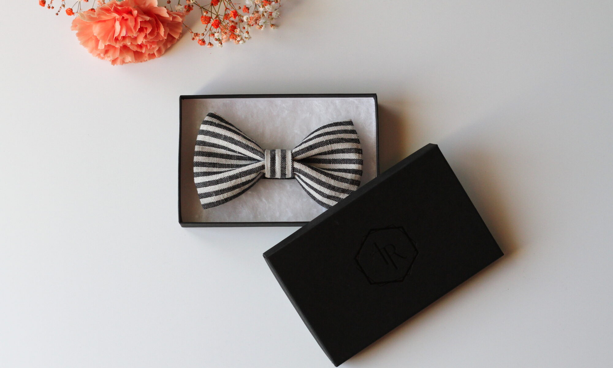 Raidallinen pellavarusetti rasiassa, vieressä nojaa rasiaa vasten musta kansi ja yläreunassa oranssi kukka koristeena.