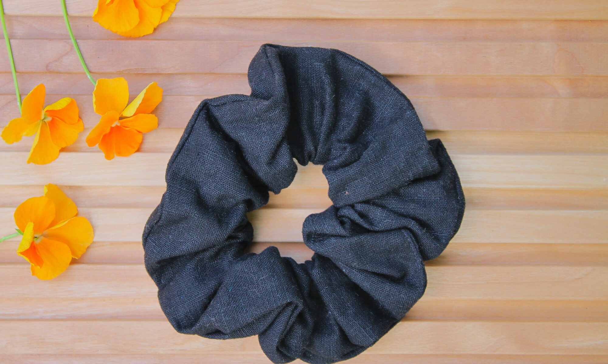 Pellavainen hiusdonitsi mustan sävyssä puupintaisella taustalla, vieressä oransseja kukkia.