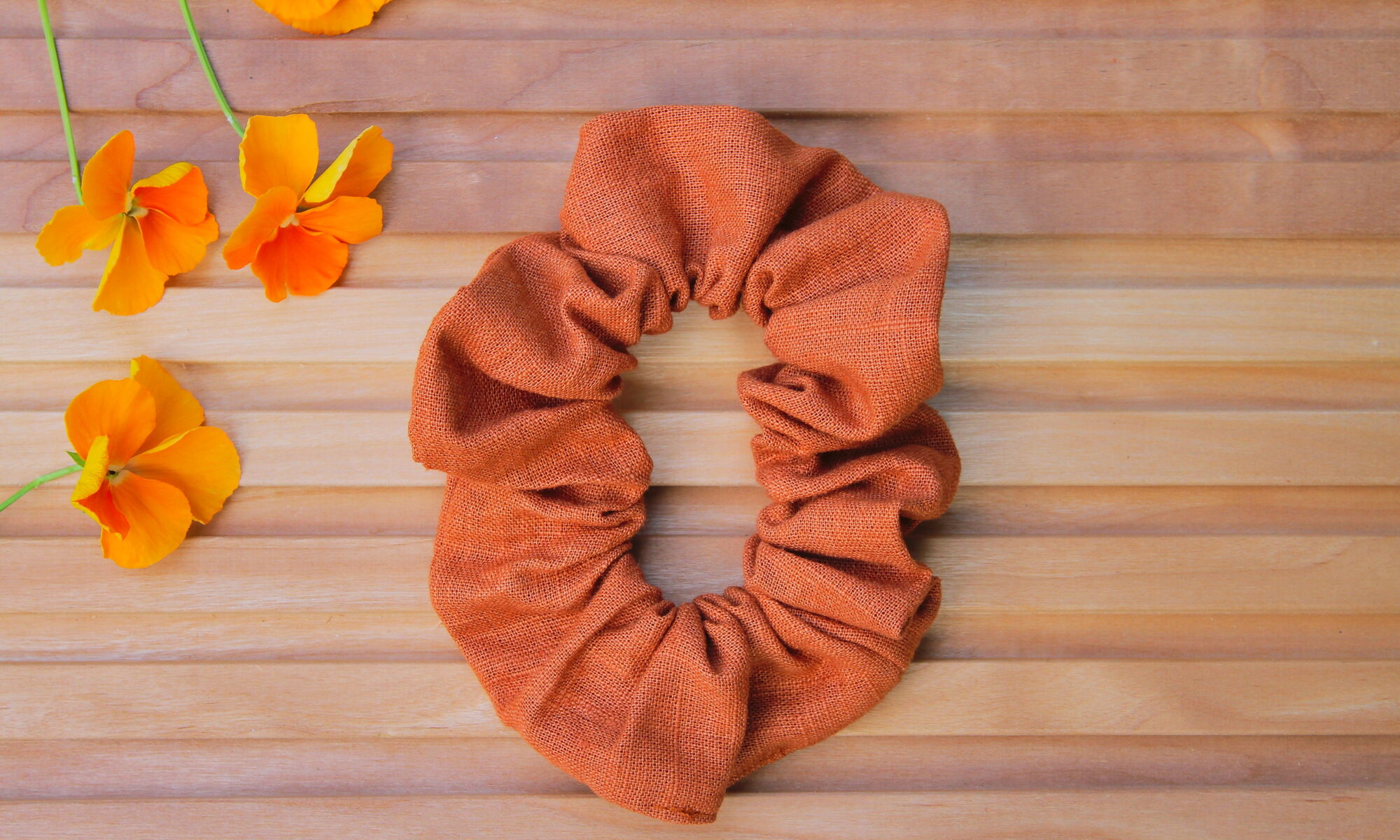 Pellavainen hiusdonitsi lämpimän kanelin sävyssä puupintaisella taustalla, vieressä oransseja orvokkeja.