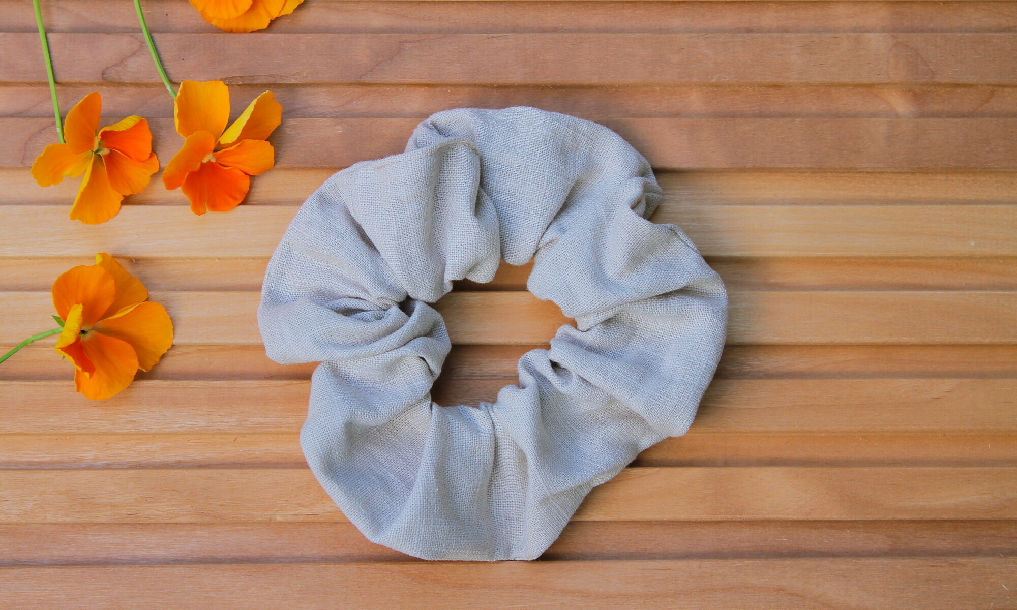 Pellavainen hiusdonitsi vaalean hiekan sävyssä puupintaisella taustalla, vieressä oransseja orvokkeja.