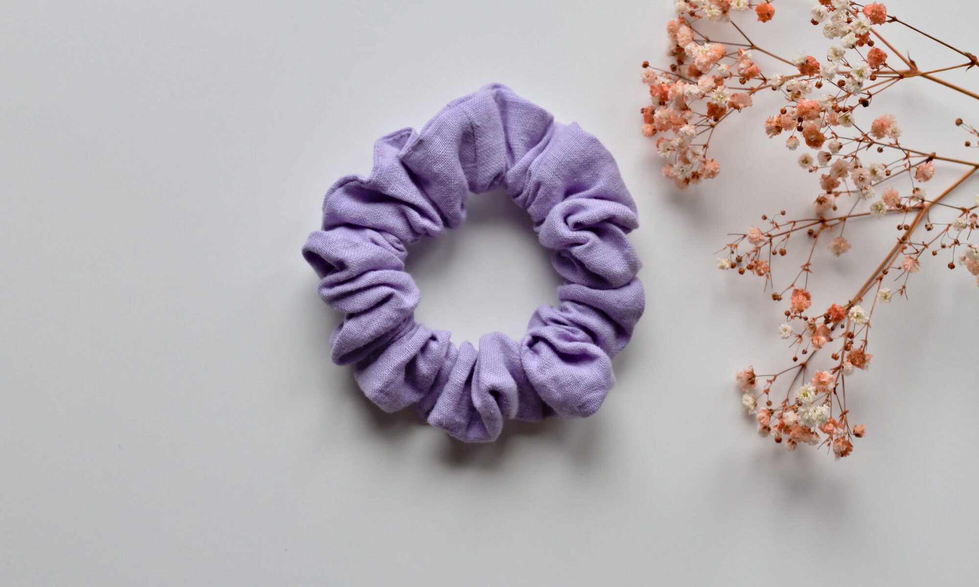 Kuvassa valkoista taustaa vasten pellavainen vaalean liilan sävyinen hiusdonitsi. Taustalla kuivakukkia.